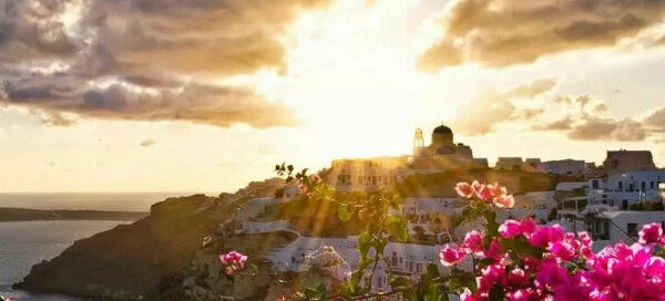 Sun setting behind the a cliff with pink flowers and white-washed houses as captured in a Santorini photo tour